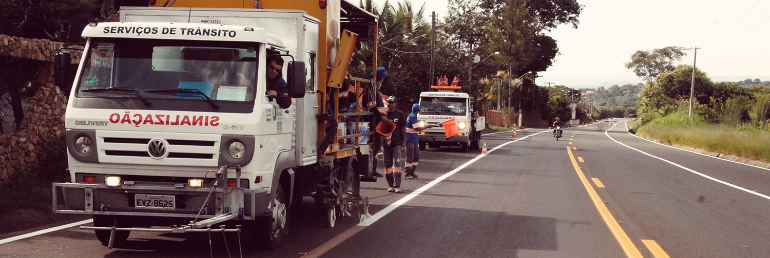 Na foto do banner, caminhões de serviço da EMDEF estão estacionados a margem de uma via enquanto funcionários sinalizam com cones de sinalização o local onde será realizado serviços de manutenção.